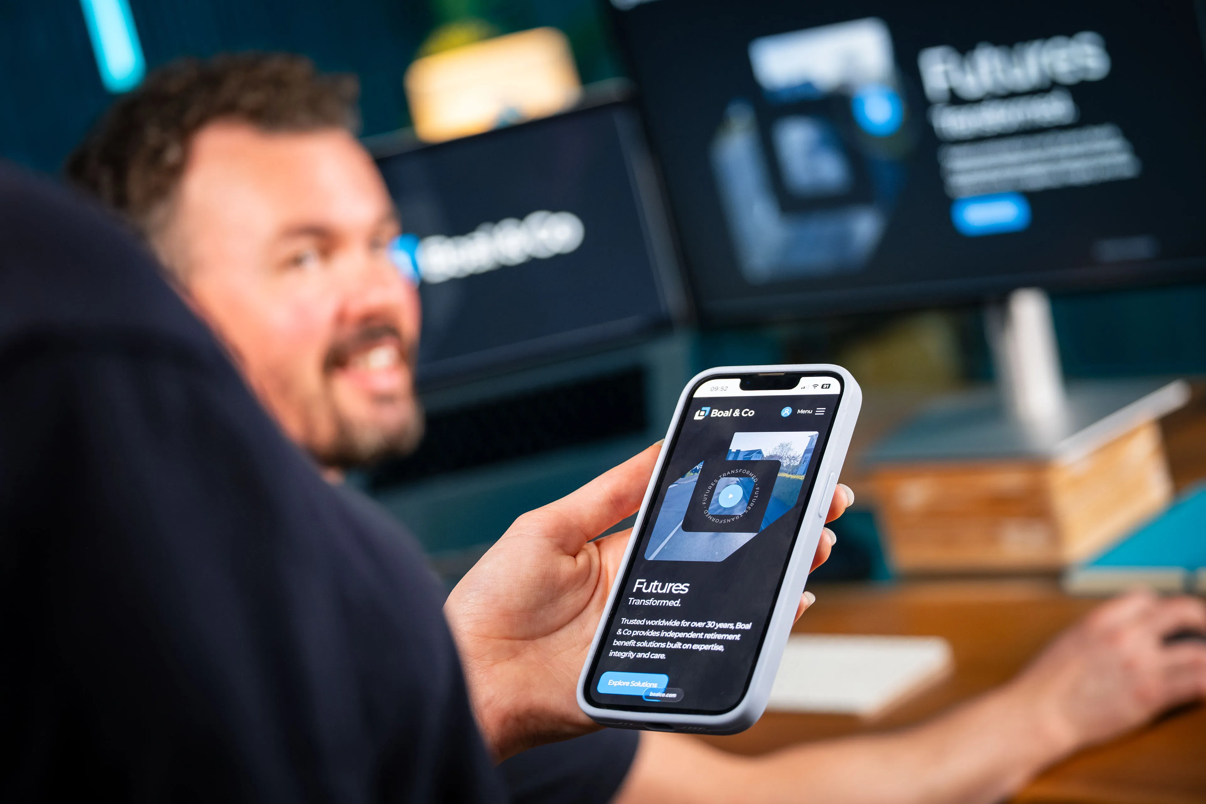 A hand holds a phone displaying the Boal & Co "Futures Transformed" homepage while a smiling Adrian Crellin looks on out of focus, in front of monitors showing the same design, at the DotPerformance office.